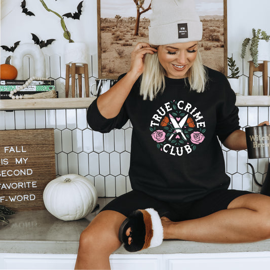 A woman in a cozy "True Crime Junkie" sweatshirt by badd-boss-f3a0 sits on a kitchen counter decorated for Halloween with pumpkins, bats, and a sign that says, "Fall is My Second Favorite F-Word." She holds a "Witch Brew" mug and smiles while adjusting her beanie, clearly embracing her True Crime Junkie spirit.