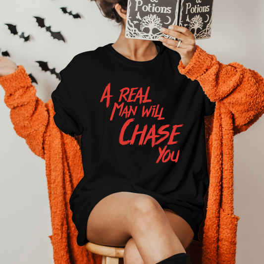 A person in a badd-boss-f3a0 oversized black "A Real Man Will Chase You" T-shirt sits on a stool, partially obscuring their face with an open book titled "Potions." They don an orange cardigan, while black bat decorations adorn the background, creating the perfect Halloween t-shirt scene.