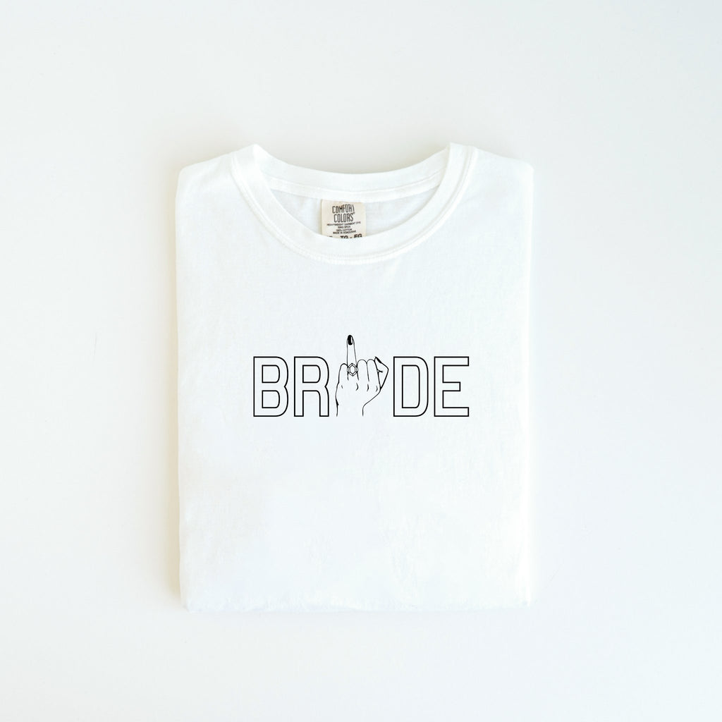 The Bride Tee from badd-boss-f3a0 offers a playful spin on bridal attire by incorporating the word "BRIDE" with the letter "I" humorously replaced by an image of a hand showing the middle finger. This unique design is perfect for adding a touch of humor to any occasion and makes an excellent wedding gift. The shirt is displayed neatly folded on a clean, white surface.