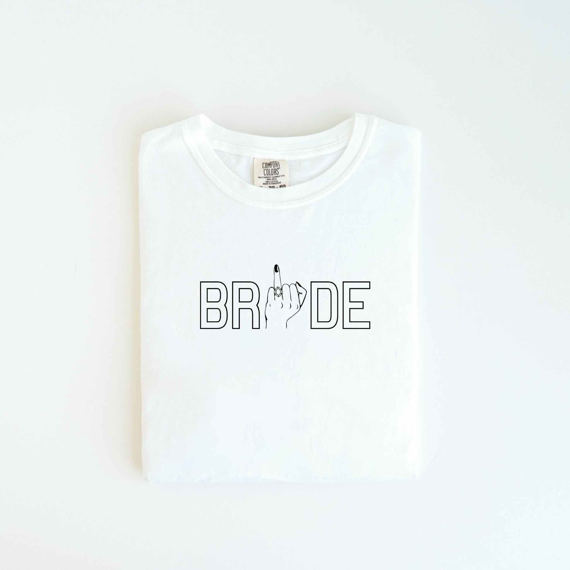 The Bride Tee from badd-boss-f3a0 offers a playful spin on bridal attire by incorporating the word "BRIDE" with the letter "I" humorously replaced by an image of a hand showing the middle finger. This unique design is perfect for adding a touch of humor to any occasion and makes an excellent wedding gift. The shirt is displayed neatly folded on a clean, white surface.