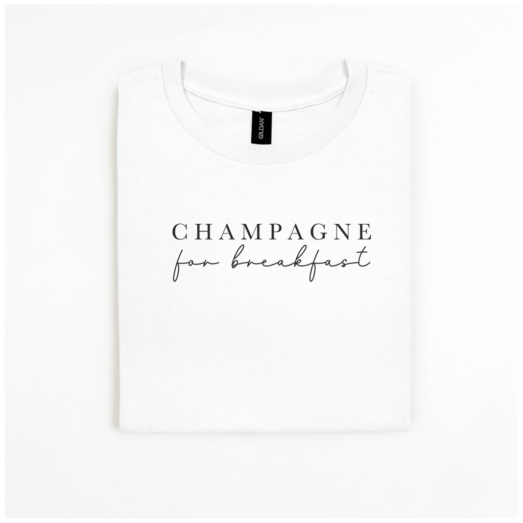 The Champagne For Breakfast Tee from badd-boss-f3a0 is displayed on a plain white background. It's a folded white Gildan Softstyle t-shirt featuring the phrase "Champagne for breakfast" in elegant black script on the front, crafted from 100% cotton to ensure a luxurious feel.