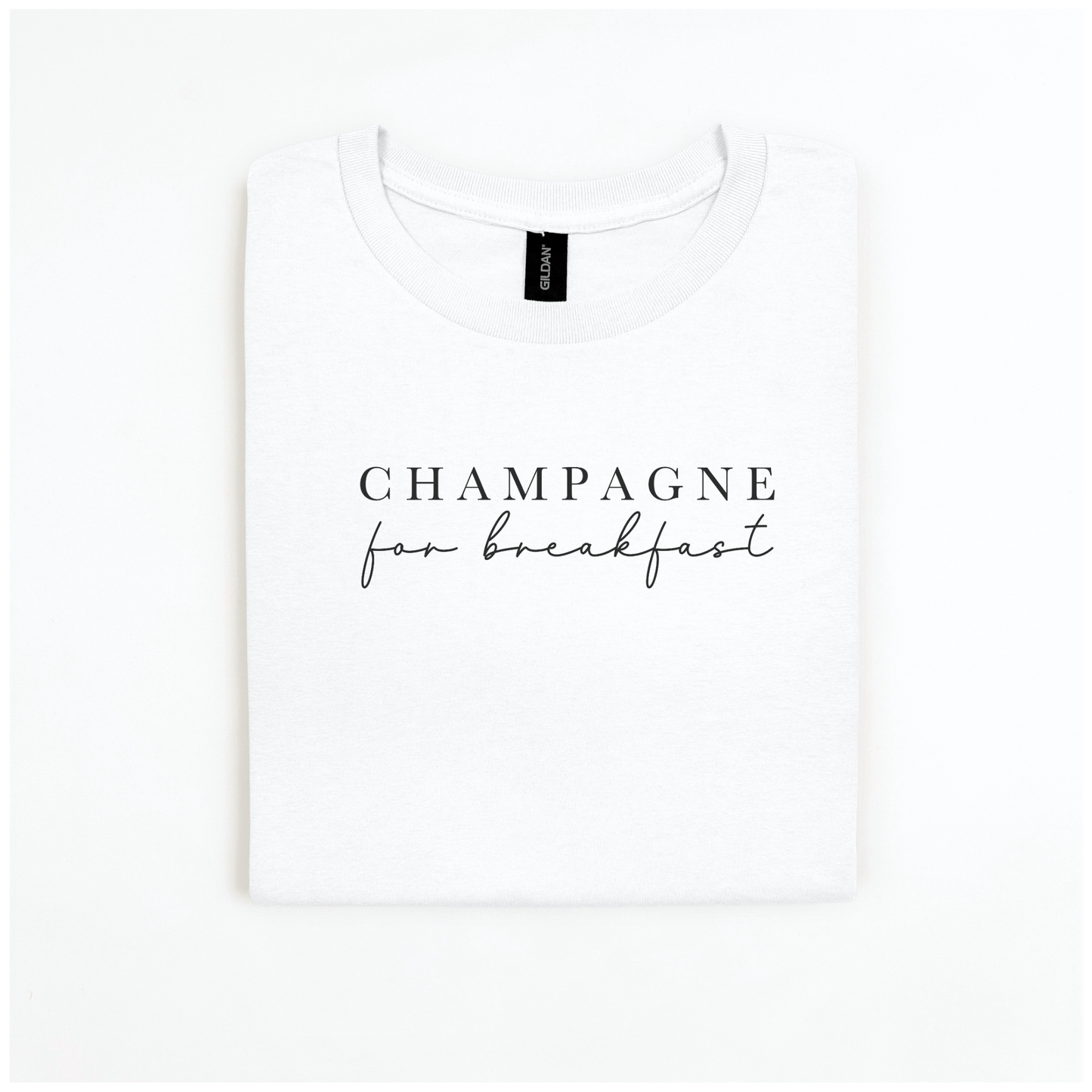 The Champagne For Breakfast Tee from badd-boss-f3a0 is displayed on a plain white background. It's a folded white Gildan Softstyle t-shirt featuring the phrase "Champagne for breakfast" in elegant black script on the front, crafted from 100% cotton to ensure a luxurious feel.