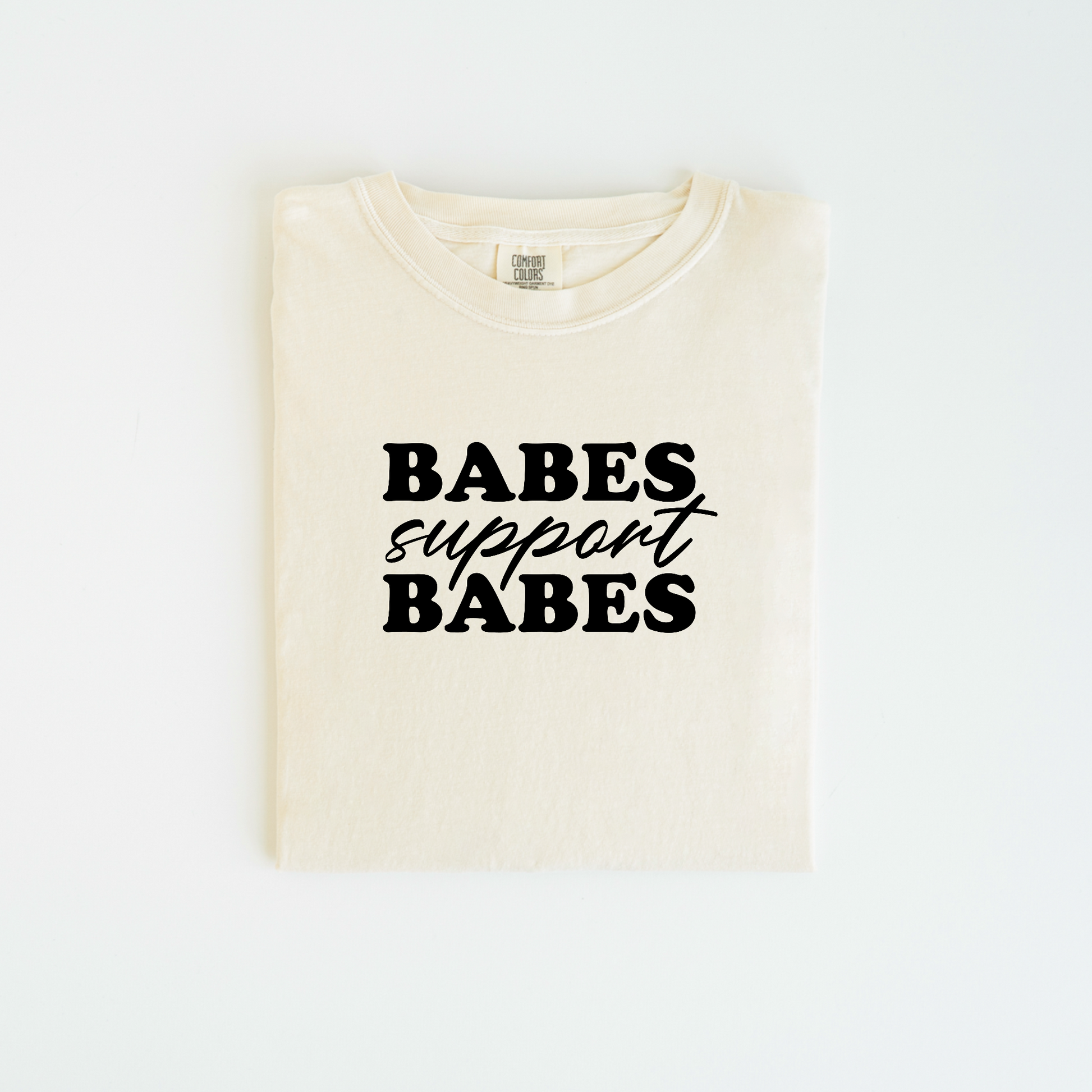A folded cream-colored "Babes Support Babes T-shirt" from badd-boss-f3a0, made of 100% cotton, features bold black lettering on the front. This unisex fit shirt is beautifully displayed against a white background.