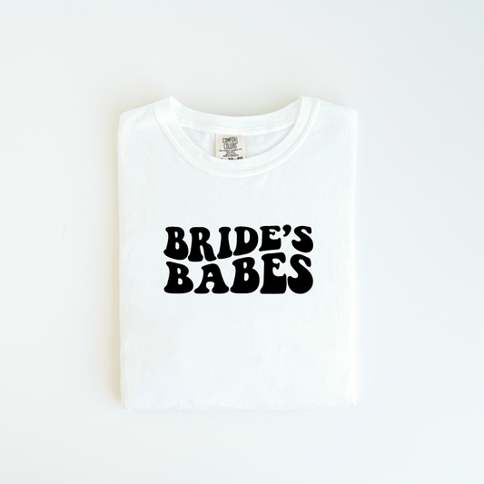 The Bride Babe Tee by badd-boss-f3a0 is folded neatly on a flat surface, highlighting its unisex fit. Made from 100% cotton, it sports bold black text that says "BRIDE'S BABES" in a playful retro font, combining style with comfort.