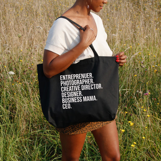 A person stands in a grassy field carrying the Badd Boss Entrepreneur Tote Bag, a durable black tote emblazoned with white text listing various roles: "ENTREPRENEUR. PHOTOGRAPHER. CREATIVE DIRECTOR. DESIGNER. BUSINESS MAMA. CEO." This custom-designed tote is perfect for showcasing talent while supporting local small businesses, and the person wears a white t-shirt and leopard print skirt.