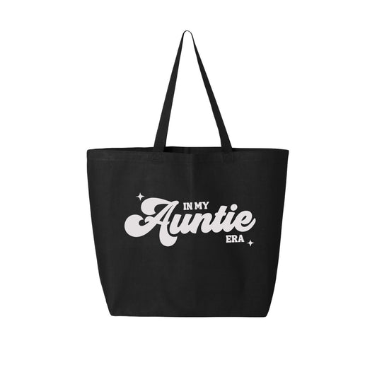 The "In My Auntie Era Tote Bag" by Badd Boss is a black tote with two shoulder straps, featuring the phrase "In My Auntie Era" in bold white cursive and block letters, adorned with small star graphics for decoration. This custom design exudes a simple, minimalist aesthetic.